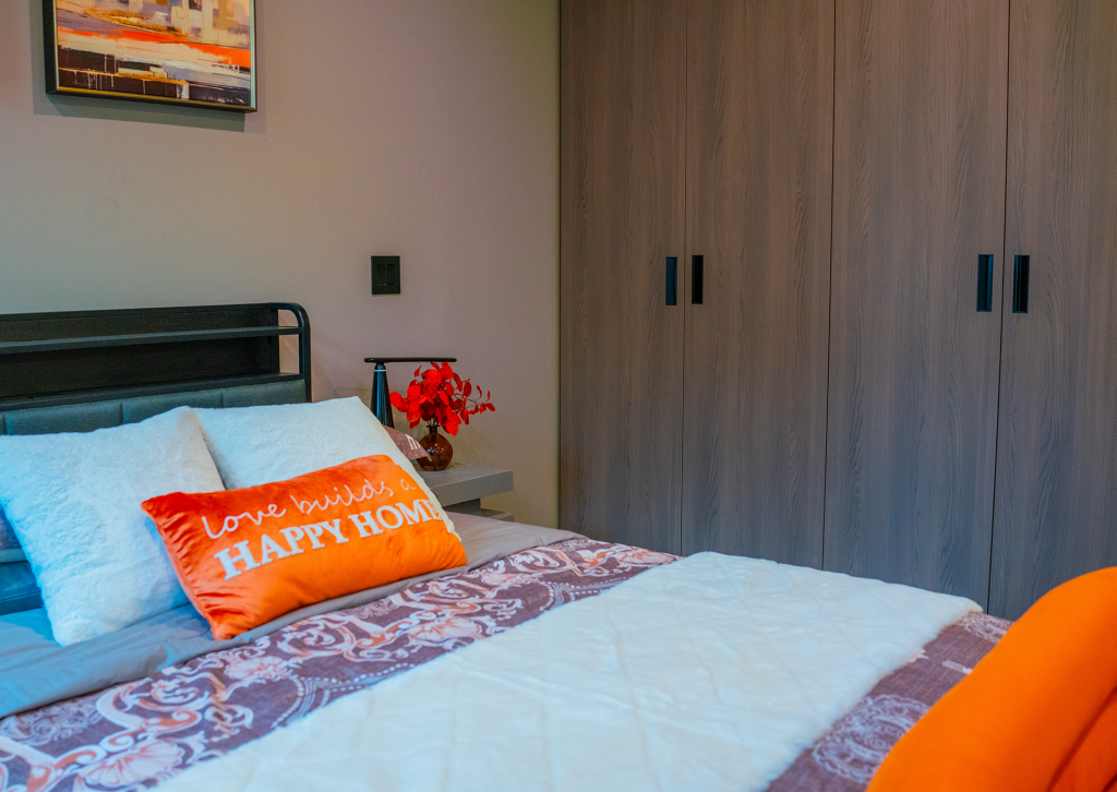 Bedroom with a wooden wardrobe, orange accent pillow reading Love Builds a Happy Home, and a nightstand with red flowers