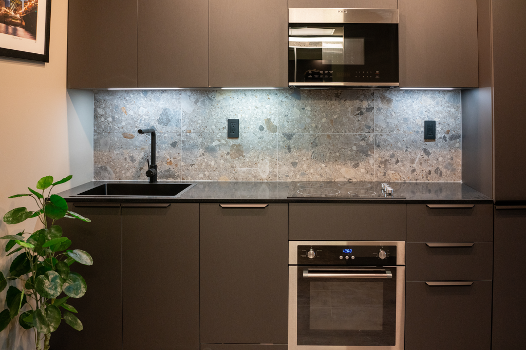 Modern kitchenette with dark cabinets, built-in oven, microwave, cooktop, sink and stone tile backsplash