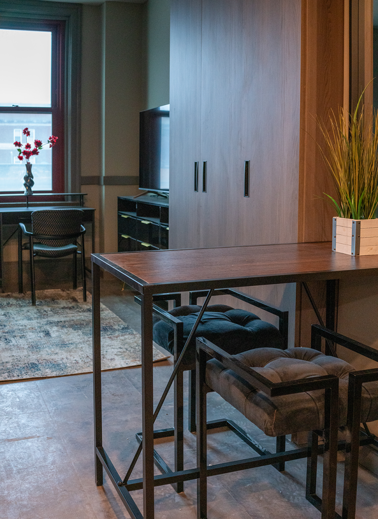 High-top table with cushioned stools sits near a wardrobe, TV console, desk, and window in an apartment.
