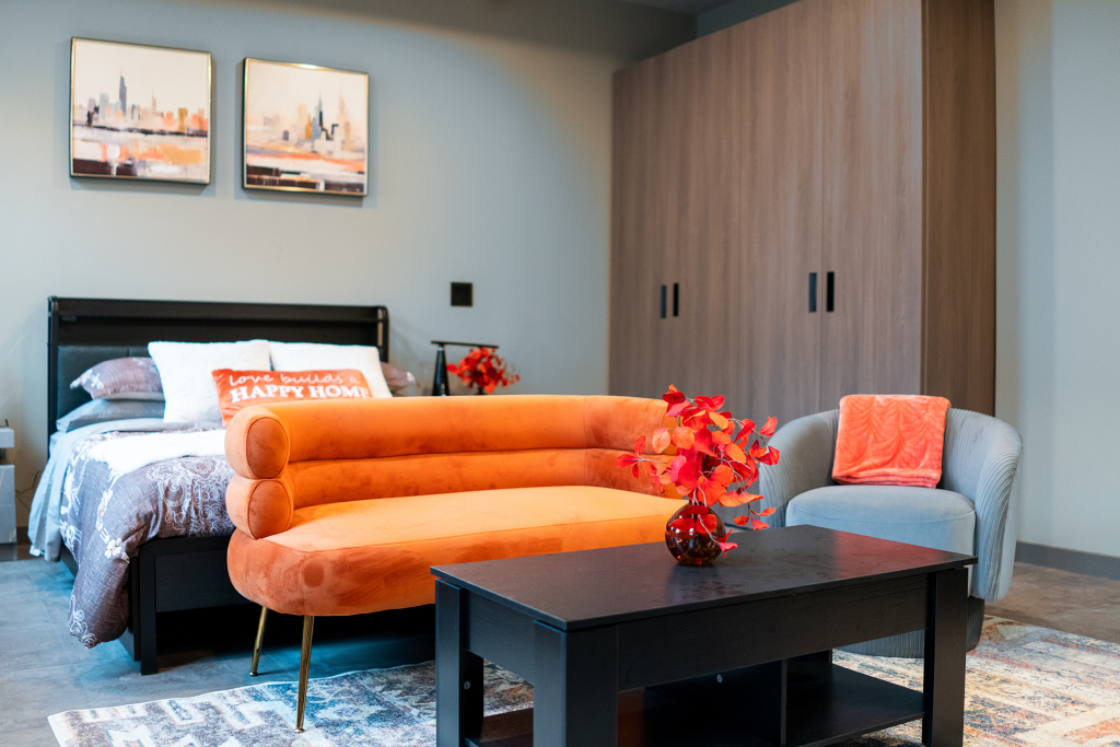 Studio apartment with orange velvet bench, gray armchair, bed, and city skyline artwork