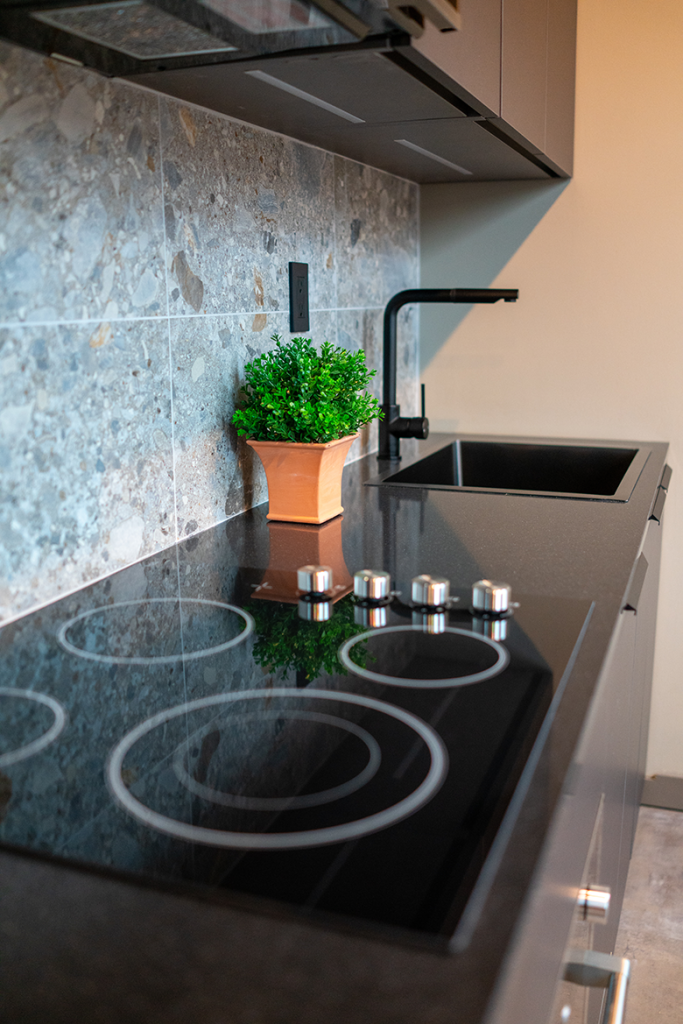 Black glass cooktop with stainless knobs beside a dark sink, black faucet, and small potted plant on a stone countertop