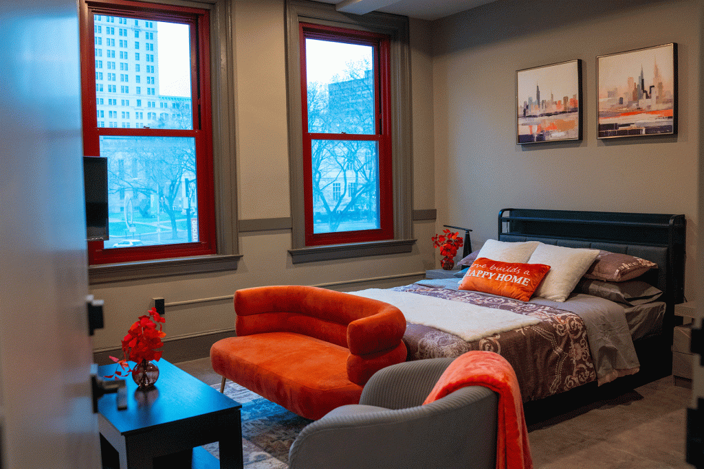 Bedroom with red-framed windows overlooking city buildings, an orange bench, and city skyline artwork above the bed