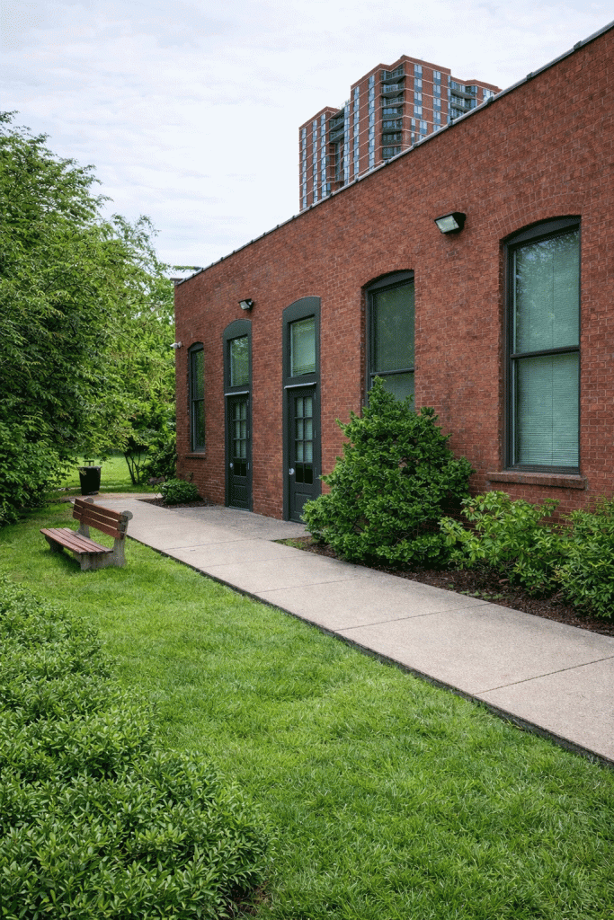 Brick building exterior with arched windows, walkway, bench, and lawn, with a high-rise visible behind