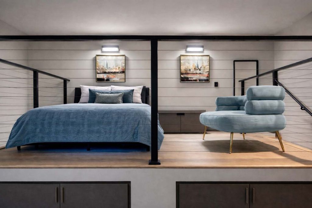 Loft bedroom with blue bedding and matching lounge chair separated by a metal railing above cabinetry